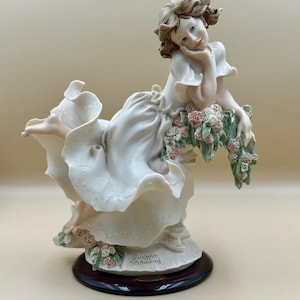 Könnte beinhalten: Porzellanfigur einer Frau in einem fließenden weißen Kleid, die sich zurücklehnt und Blumen hält. Die Figur hat einen hellbraunen Sockel und einen dunkelbraunen Holzständer. Das Haar der Frau ist hellbraun, und sie hat einen ruhigen Ausdruck.