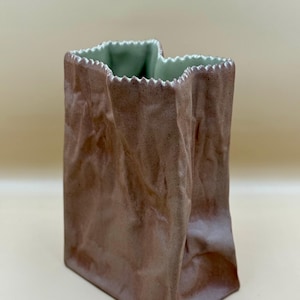 Op de afbeelding: Een bruine keramische vaas in de vorm van een papieren zak. De vaas heeft een getextureerd oppervlak met een gekreukte uitstraling. De binnenkant is lichtgroen en de rand is gekarteld.