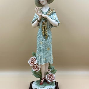 Könnte beinhalten: Porzellanfigur einer Frau in Vintage-Kleid und Hut, die eine Rose hält. Die Figur ist hellgrün mit goldenen Akzenten und steht auf einem Sockel mit Rosen und dem Wort "Rose". Der Sockel ist dunkelbraun.