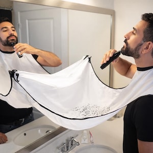 May include: A white beard bib with black trim is held up to catch beard trimmings. The person in the image is trimming their beard with an electric trimmer. The bib is suspended by hooks attached to a mirror.