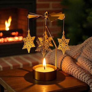 May include: A gold-colored candle holder with a spinning snowflake design. The candle holder has a round base with a lit candle, and three snowflake ornaments hang from the top. The background features a fireplace and a cozy blanket.