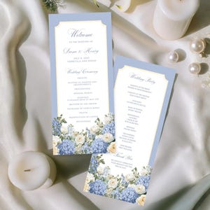 May include: Two pale blue wedding programs with floral detailing and text. The programs are adorned with blue and white flowers at the base. Two white candles and pearls are also visible, set against a white satin backdrop.
