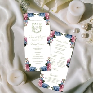 以下が含まれることがあります： 青とピンクのアジサイの花柄が特徴のウェディングステーショナリー。招待状には「Clara & Edward」の名前と結婚式の詳細が記載されています。2本の白いキャンドル、真珠、花が白いサテンの表面に配置されています。