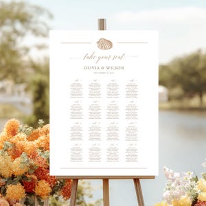 Könnte beinhalten: Ein weißes Sitzplan mit goldenem Text und einer Muschel-Illustration. Der Text lautet "take your seat" über den Namen Olivia & Wilson und dem Datum 31. Dezember 2021. Die Tafel steht auf einer Holzstaffelei.