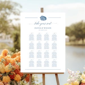 May include: A white seating chart on a wooden easel, featuring a navy blue seashell graphic and the words "take your seat." The names Olivia & Wilson and the date December 31, 2021, are also displayed. The chart lists guest names by table.