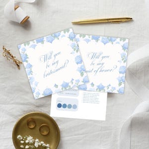 以下が含まれることがあります： 青いアジサイの花柄の縁取りと「Will you be my bridesmaid?」と「Will you be my maid of honor?」の文字が入った2枚の招待状。金のペン、2つの指輪が入った小さな金の皿、カラーパレットカードも写っています。