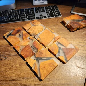 Op de afbeelding: Zes vierkante onderzetters gemaakt van hout en helder hars. De onderzetters hebben een natuurlijk houtnerfpatroon met zwarte harswervelingen.