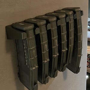 May include: A black wall-mounted magazine holder with seven magazines inserted. The holder is made of plastic and has a textured surface. The magazines are all black and have a similar design.