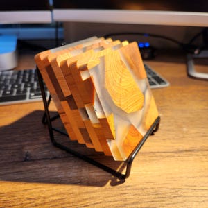 Op de afbeelding: Set van vier houten onderzetters met een transparante epoxyhars top. De onderzetters zijn gestapeld en worden getoond op een zwarte metalen standaard.