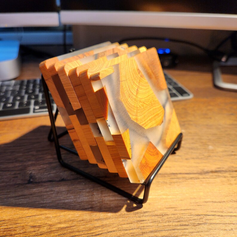 Op de afbeelding: Set van vier houten onderzetters met een transparante epoxyhars top. De onderzetters zijn gestapeld en worden getoond op een zwarte metalen standaard.