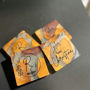 Op de afbeelding: Vier vierkante onderzetters gemaakt van hout en hars met een heldere epoxy-afwerking. De onderzetters hebben een natuurlijk houtnerfpatroon met een grijs en zwart marmerpatroon. Een onderzetter heeft de letters "R" en de datum "08.17.24" erop gegraveerd. Een andere onderzetter heeft de namen "Tyler" and "Christina" erop gegraveerd. De onderzetters zijn perfect om elke woning een vleugje rustieke elegantie te geven.