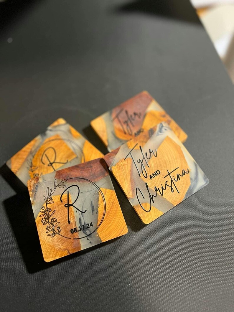 Op de afbeelding: Vier vierkante onderzetters gemaakt van hout en hars met een heldere epoxy-afwerking. De onderzetters hebben een natuurlijk houtnerfpatroon met een grijs en zwart marmerpatroon. Een onderzetter heeft de letters "R" en de datum "08.17.24" erop gegraveerd. Een andere onderzetter heeft de namen "Tyler" and "Christina" erop gegraveerd. De onderzetters zijn perfect om elke woning een vleugje rustieke elegantie te geven.