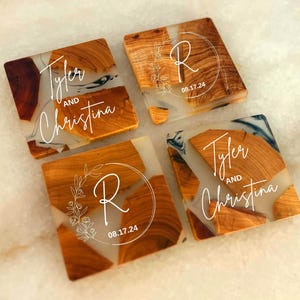Op de afbeelding: Vier vierkante houten onderzetters met een transparante epoxyhars top. De onderzetters hebben een wit swirl-ontwerp en de namen "Tyler en Christina" en de letter "R" met de datum "08.17.24".