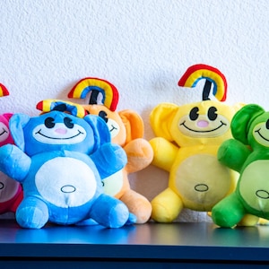 May include: A collection of plush toys in pink, blue, orange, yellow, and green. Each toy has a smiling face, a white belly, and a rainbow on top. The toys are arranged on a dark blue surface against a white wall.