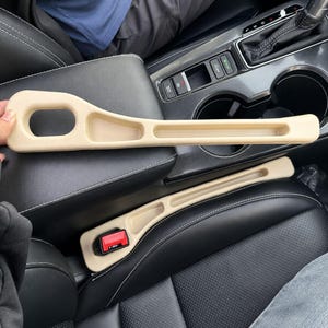 May include: A beige plastic car seat gap filler organizer featuring a long, narrow tray design with a cutout handle section. The accessory is shaped to fit between the center console and the seat, providing a storage compartment for small items.