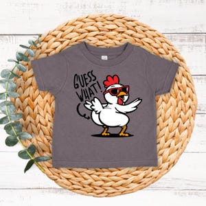 May include: A dark gray short-sleeved t-shirt with a white chicken wearing sunglasses and the text "Guess What!" in red.