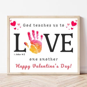 May include: A framed print with the word "LOVE" in large black letters, featuring a handprint in shades of pink, orange, and yellow. The text "God teaches us to" and "one another" are above and below the word "LOVE", with "Happy Valentine's Day!" at the bottom.