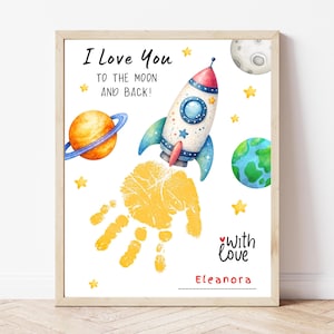 May include: A framed print with a space theme. It features a rocket ship, planets, and stars, with the text "I Love You to the Moon and Back!" and a handprint. The print is in a wooden frame.