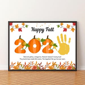 Puede incluir: Una impresión enmarcada con un borde negro presenta las palabras "Happy Fall" sobre los números naranjas "202" diseñados como calabazas. Una huella de mano amarilla y hojas de otoño decoran el fondo blanco. El texto dice: "Rojo y amarillo, naranja también. ¡Hojas de otoño en cada tono! Manos pequeñas han hecho esta escena. El otoño más feliz que hayas visto."