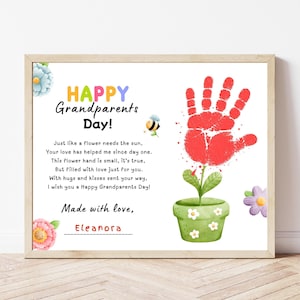 May include: A framed print for Grandparents Day featuring a red handprint flower in a green pot, a small bee, and colorful flowers. The text reads: Happy Grandparents Day! Just like a flower needs the sun, Your love has helped me since day one. Made with love, Eleanora.
