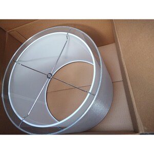 Op de afbeelding: Een cilindrische lampenkap met een zilverkleurige, getextureerde buitenkant en een witte binnenkant. De lampenkap heeft een metalen frame en staat in een kartonnen doos.