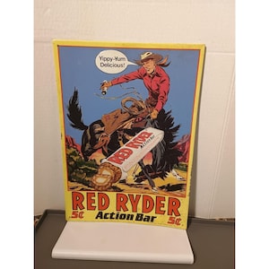 May include: A vintage Red Ryder Action Bar sign with a cowboy on a bucking bronco. The sign has a yellow border and includes the text "Yippy-Yum Delicious!" in a speech bubble. The candy bar is shown in the center of the sign.