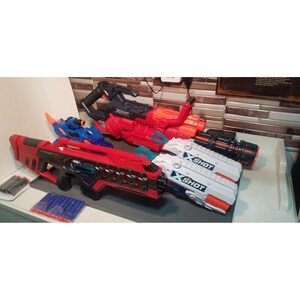 May include: A collection of toy blasters in various colors, including red, black, white, and blue. The blasters have the "X-SHOT" logo and come with foam darts. The toys are designed for children's play and are arranged on a table.