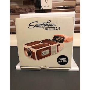 以下が含まれることがあります： 茶色とクリーム色のデザインのスマートフォンプロジェクター2.0ボックス。「Smartphone Projector 2.0」と「Cinema in a Box！」の文字が入っています。プロジェクターは茶色とシルバーのデザインです。