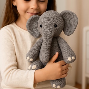 May include: A grey crochet elephant toy with large ears, a trunk, and small black eyes. The elephant has white accents on its feet and is being held by a person wearing a cream-coloured sweater. The elephant is a handmade toy.