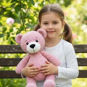 May include: A pink crochet teddy bear with a white snout and black eyes. The bear is held by a person wearing a white long-sleeved top. The background is a blurred outdoor scene with green leaves and branches.