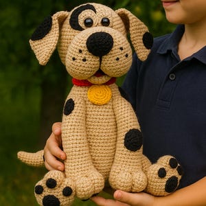 Puede incluir: Un peluche de perro hecho a mano con ganchillo en color beige con manchas negras, orejas caídas y una nariz negra. Tiene un collar rojo con una etiqueta amarilla. El perro está siendo sostenido por una persona. El peluche mide unos 30 cm de alto.