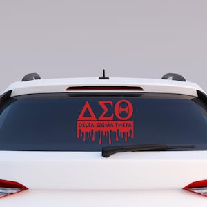 以下が含まれることがあります： 白い車のリアウィンドウに貼られた、赤いビニール製のステッカー。ステッカーには「&Delta;&Sigma;&Theta;」の文字と「DELTA SIGMA THETA」の文字が書かれており、文字が滴り落ちているように見える。