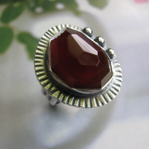 May include: A silver ring with a red carnelian gemstone set in a textured bezel. The ring has a decorative, textured band.