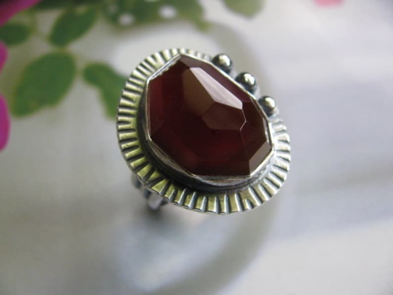 May include: A silver ring with a red carnelian gemstone set in a textured bezel. The ring has a decorative, textured band.