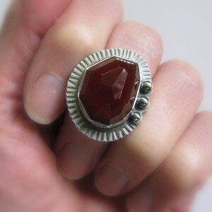 May include: A silver ring with a large, faceted, red carnelian gemstone set in a textured, geometric bezel. The ring has a decorative, textured band.