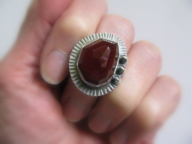 May include: A silver ring with a large, faceted, red carnelian gemstone set in a textured, geometric bezel. The ring has a decorative, textured band.