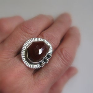 May include: A silver ring with a large, faceted, reddish-brown gemstone set in a textured, geometric bezel.