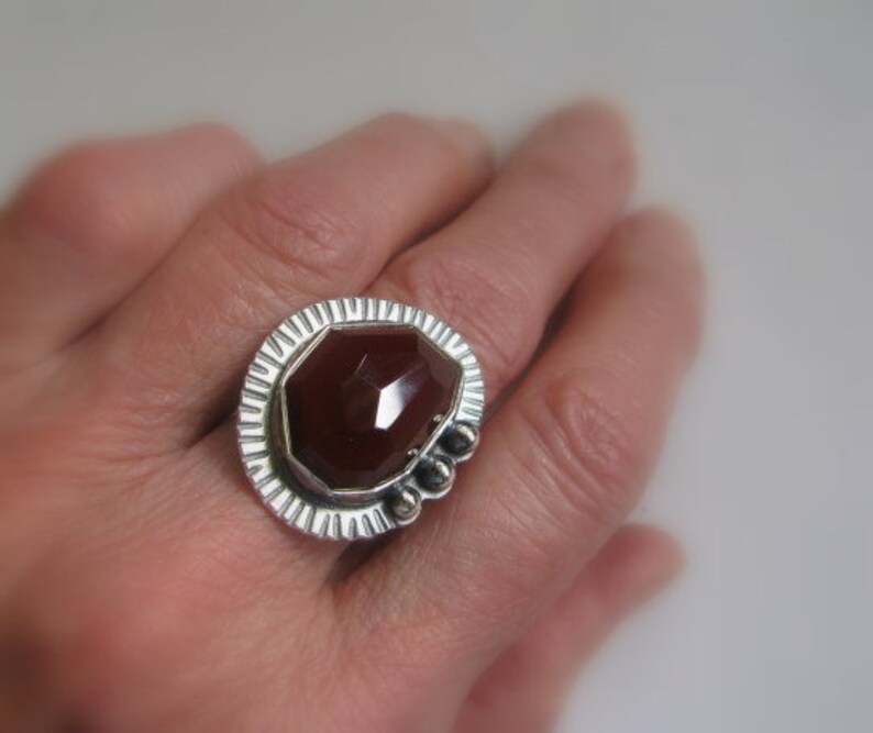 May include: A silver ring with a large, faceted, reddish-brown gemstone set in a textured, geometric bezel.