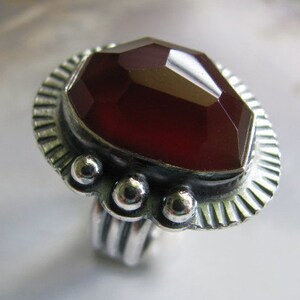 May include: A silver ring with a large, faceted, dark red gemstone set in a decorative bezel. The ring band is textured and has a series of small, round accents.