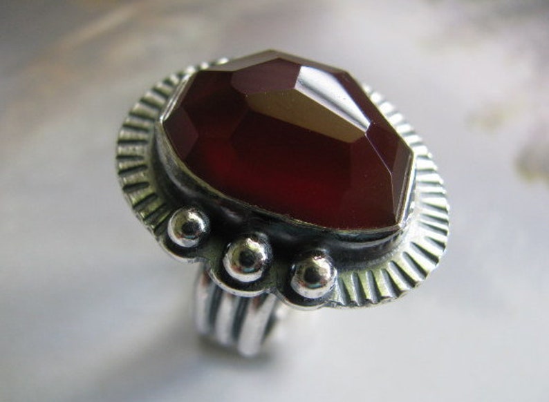 May include: A silver ring with a large, faceted, dark red gemstone set in a decorative bezel. The ring band is textured and has a series of small, round accents.
