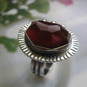 May include: A silver ring with a large, faceted, dark red gemstone set in a textured silver bezel. The ring has a wide band with a textured design.