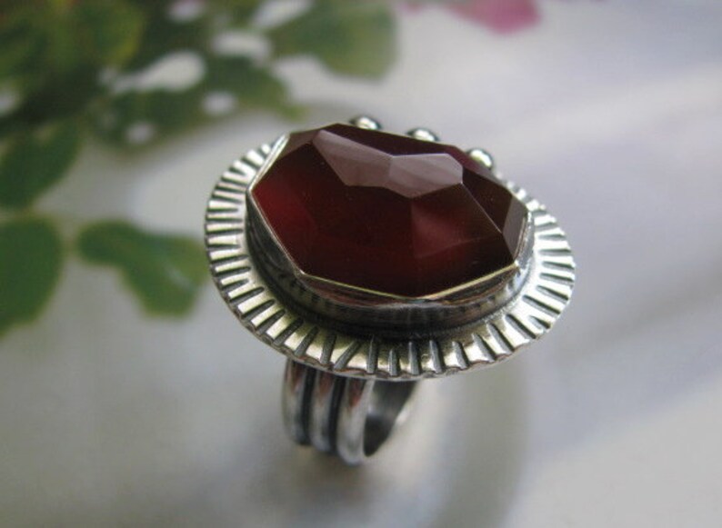 May include: A silver ring with a large, faceted, dark red gemstone set in a textured silver bezel. The ring has a wide band with a textured design.