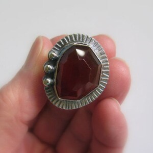 May include: A silver ring with a red gemstone set in a geometric design. The ring has a textured band and three small silver balls on the side.