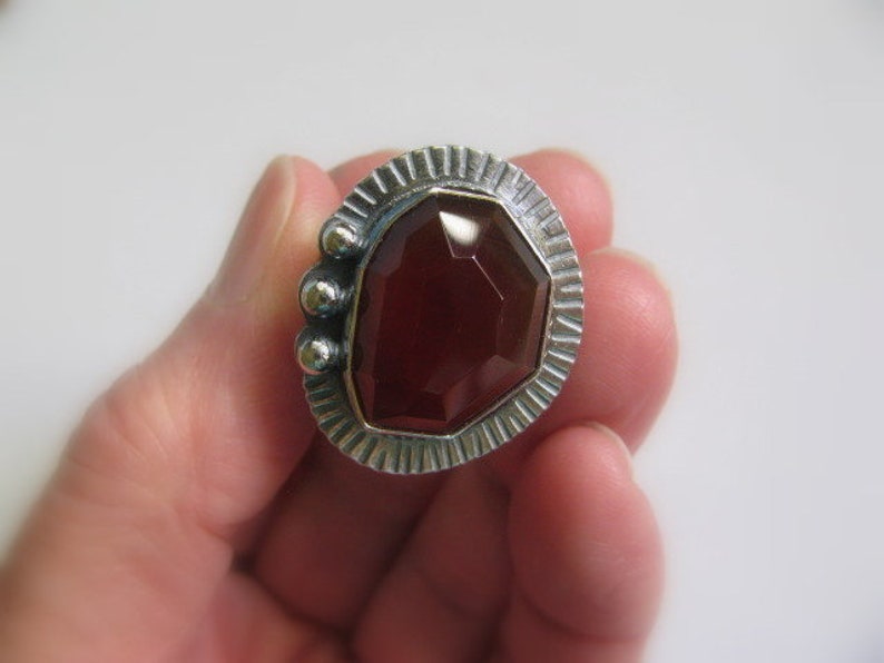 May include: A silver ring with a red gemstone set in a geometric design. The ring has a textured band and three small silver balls on the side.