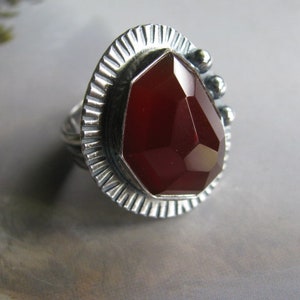 May include: A silver ring with a red gemstone set in a geometric design. The ring has a textured band and a raised setting.