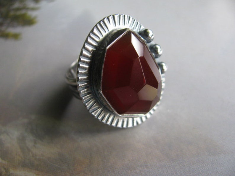 May include: A silver ring with a red gemstone set in a geometric design. The ring has a textured band and a raised setting.