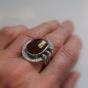 May include: A silver ring with a large, faceted, reddish-brown gemstone set in a decorative bezel. The ring has a textured band and a row of small silver beads.