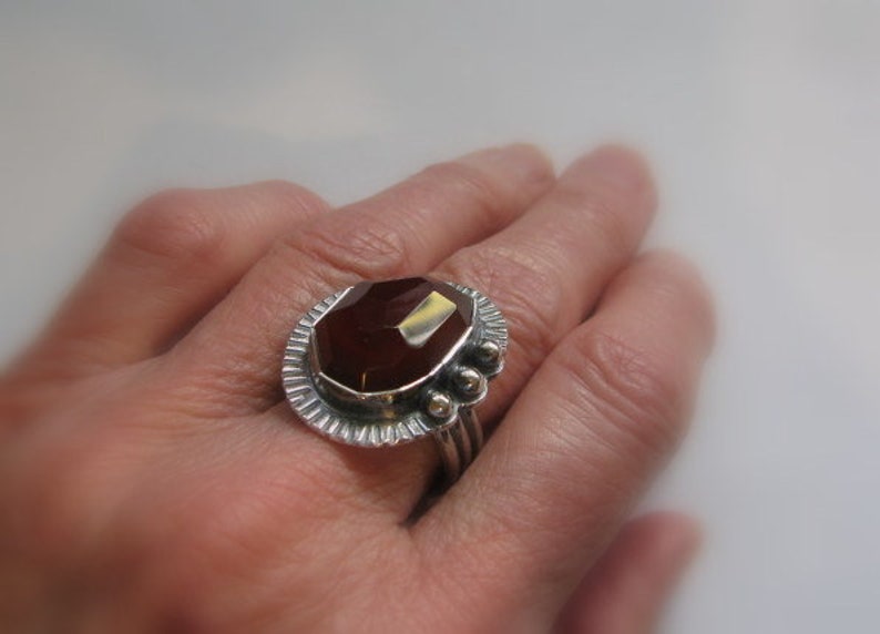 May include: A silver ring with a large, faceted, reddish-brown gemstone set in a decorative bezel. The ring has a textured band and a row of small silver beads.