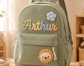 Può includere: Uno zaino in velluto a coste verde salvia con il nome Arthur ricamato in lettere di filato multicolore. La borsa presenta un'applicazione a fiore in peluche azzurro e una toppa con muso di leone sulla tasca frontale con una nappa sulla zip.