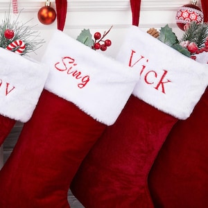 Può includere: Quattro calze di Natale rosse con polsini in finta pelliccia bianca, ciascuna ricamata con un nome in rosso. Le calze sono decorate con verde festivo e ornamenti, appese su uno sfondo bianco.